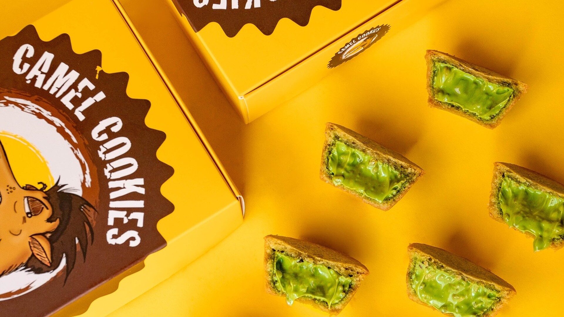 Camel Cookies boxes in yellow and chocolate brown beside stuffed cookies split open to show a bright pistachio-green centre, shot on a saffron-yellow backdrop