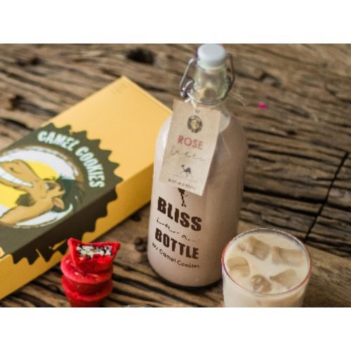 A corked glass Bliss Bottle of rose latte with a handwritten tag, on weathered wood beside a yellow Camel Cookies box, a red velvet cookie and a glass of iced latte